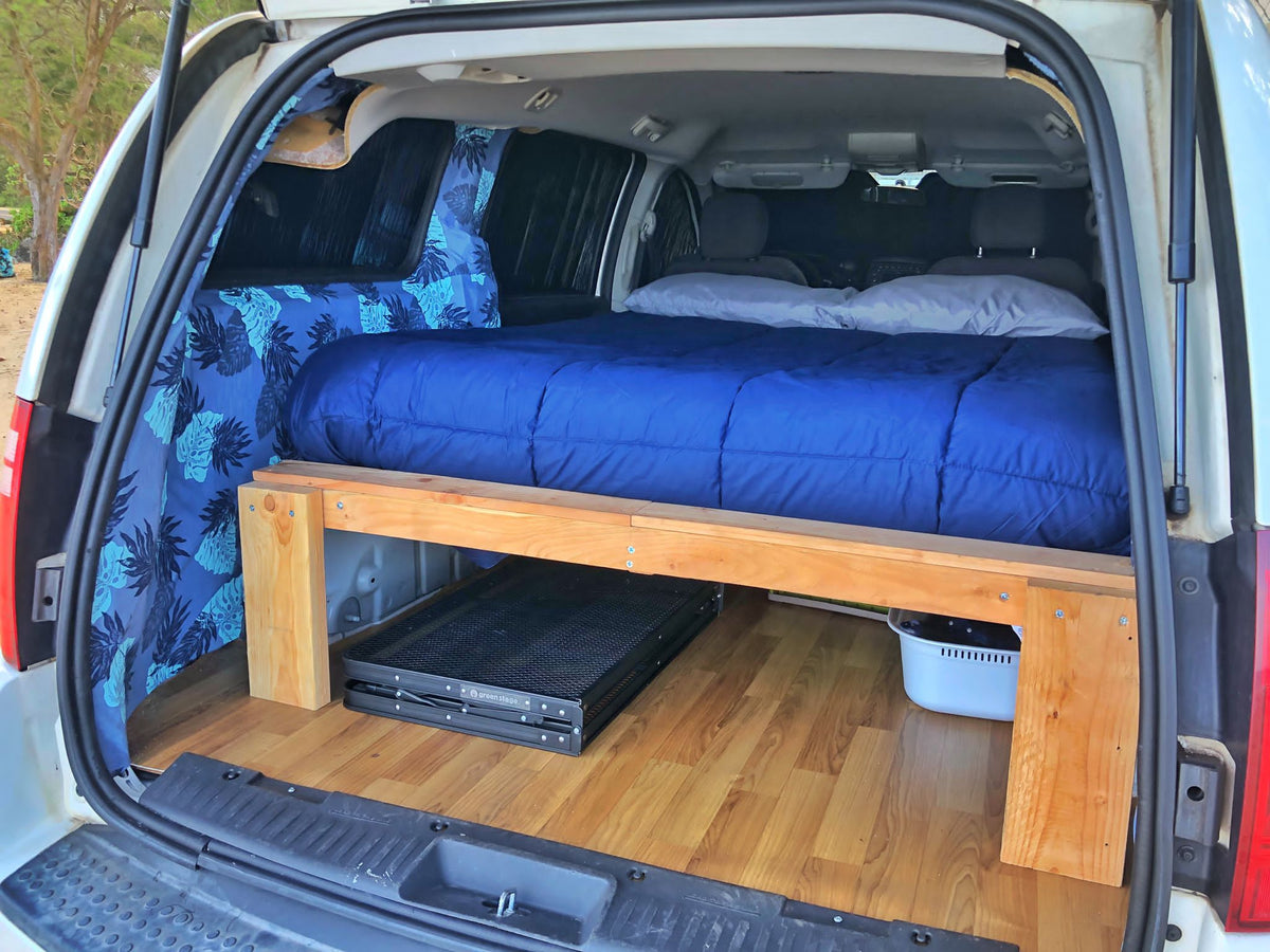 This RV rental shows a cozy van conversion: a bed with blue bedding, under-bed storage, & tropical curtains. — photo 4