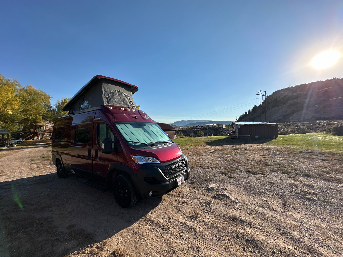 A vibrant red pop-top camper van, perfect for an RV rental, parked on a sunny dirt lot with hills & buildings.