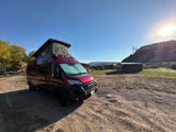 A vibrant red pop-top camper van, perfect for an RV rental, parked on a sunny dirt lot with hills & buildings.