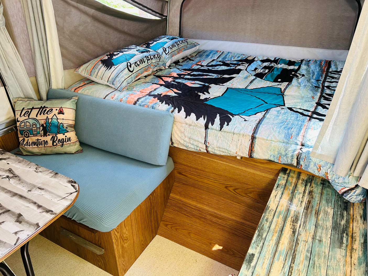 Cozy rv rental interior: bed with 'Camping' pillows, blue cushioned seating, & 'Adventure' pillow. Ready for your trip!