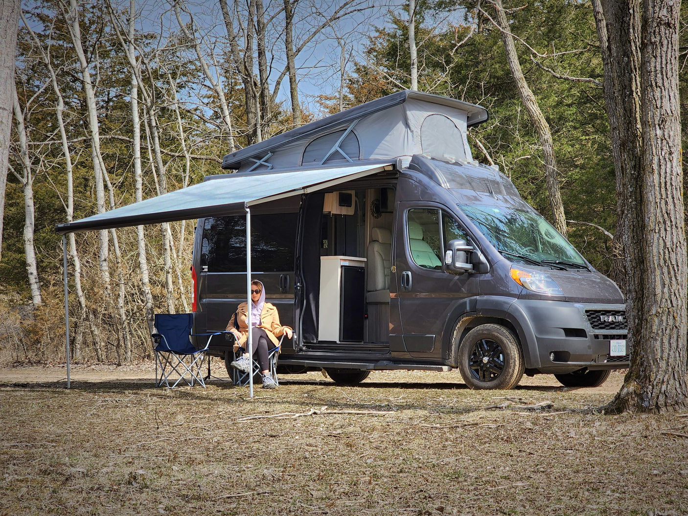 An RV rental: Woman relaxing in a folding chair under the extended awning of a grey camper van with a pop-top roof at a campsite.