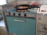Cooking in a retro RV rental kitchen: food simmering on a teal Magic Chef stove next to a sink.