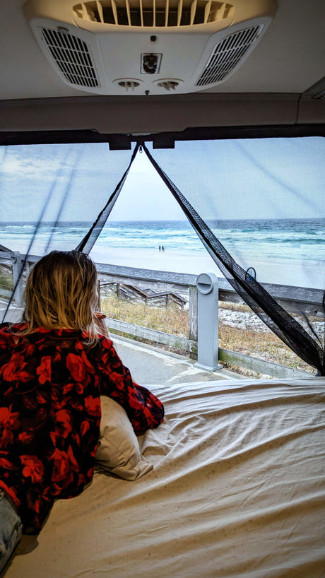 RV rental: A person lies on a bed, looking out a screened window at a tranquil beach with white sand and blue waves.