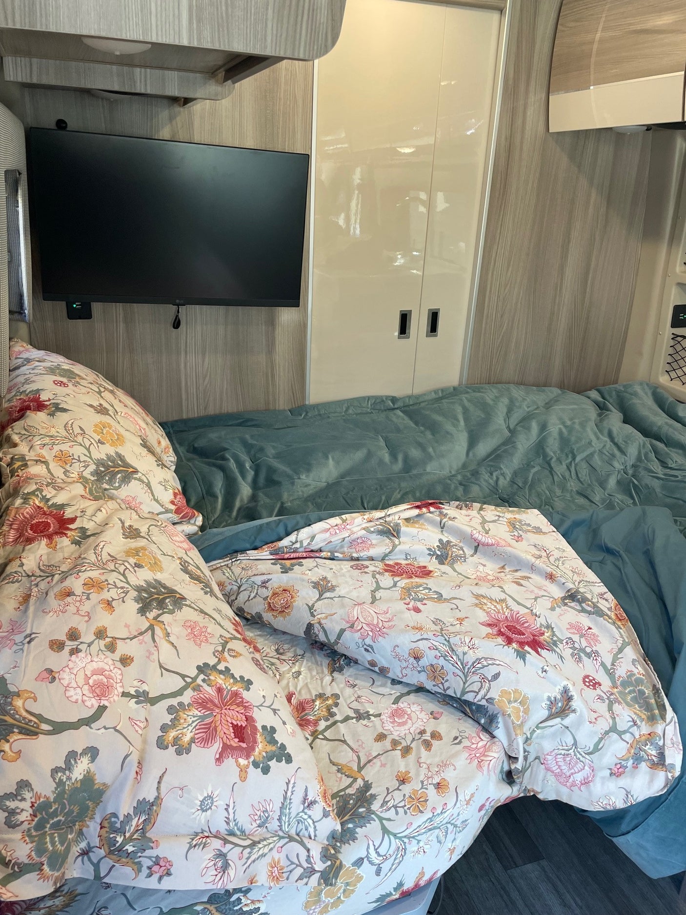 Cozy RV rental bedroom with floral bedding, wall-mounted TV, and light wood interior.