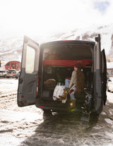 Woman in a cozy RV rental campervan, lacing boots before snowboarding at a snow-covered mountain resort. Winter adventure awaits!