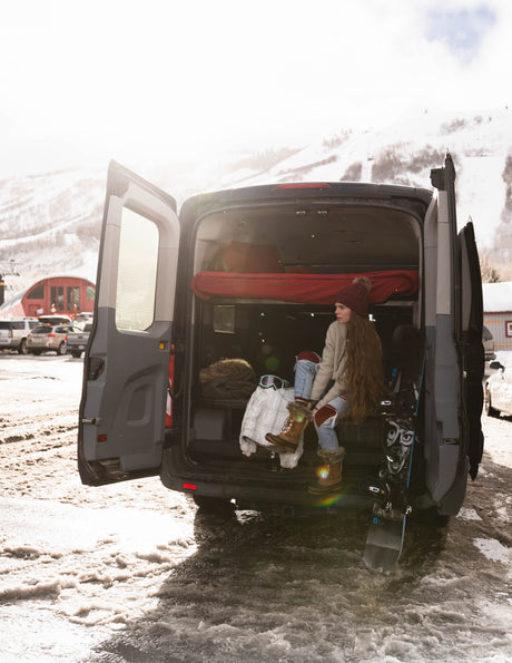 Woman in a cozy RV rental campervan, lacing boots before snowboarding at a snow-covered mountain resort. Winter adventure awaits!