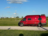 Red Solis camper van for rv rental, parked on a gravel road alongside green fields with distant farm buildings under a blue sky.