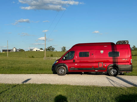 Red Solis camper van for rv rental, parked on a gravel road alongside green fields with distant farm buildings under a blue sky.