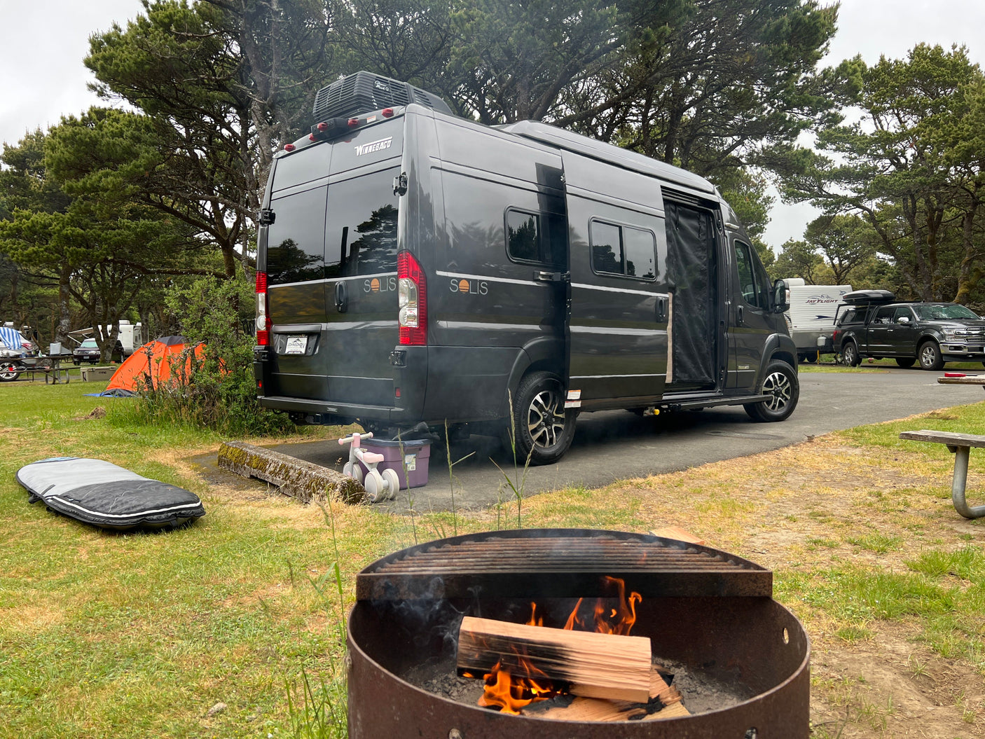 Enjoy an RV rental camping experience! A Winnebago Solis at a tree-lined campsite with a burning fire pit.