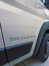 The side of a Dave & Matt Vans RV rental van, featuring the company's silver emblem on its light-colored body.