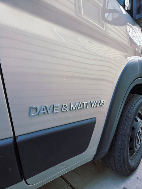 The side of a Dave & Matt Vans RV rental van, featuring the company's silver emblem on its light-colored body.