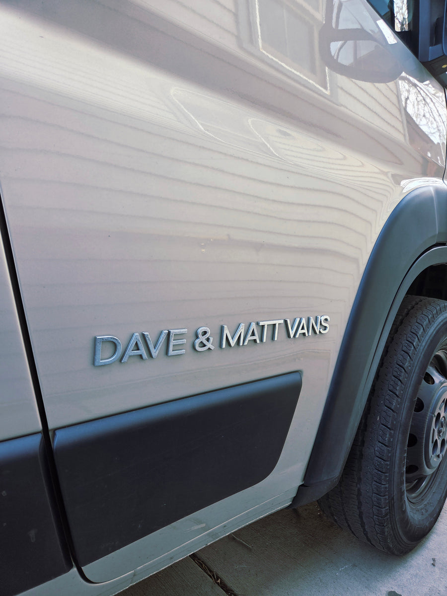 The side of a Dave & Matt Vans RV rental van, featuring the company's silver emblem on its light-colored body.