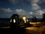 Experience an RV rental by the sea at night. Bright interior lights illuminate the van and grass under a cloudy, star-filled sky.