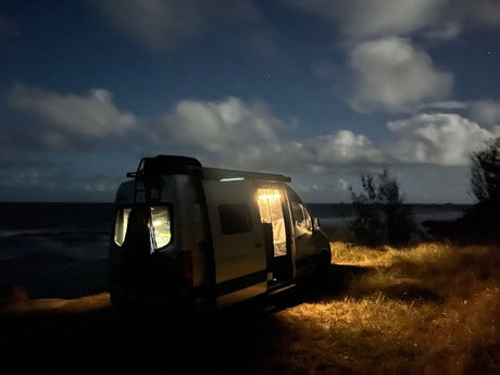 Experience an RV rental by the sea at night. Bright interior lights illuminate the van and grass under a cloudy, star-filled sky.
