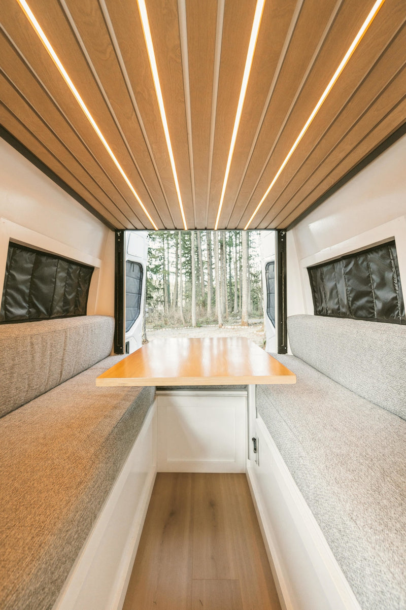 RV rental interior features: wood ceiling with linear LEDs, comfy grey benches, and a dining table. Rear doors reveal a serene forest view. — photo 3