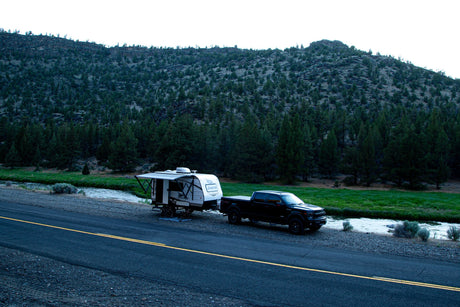 An RV rental Jayco trailer and black truck parked roadside with an extended awning by a river & wooded mountain.