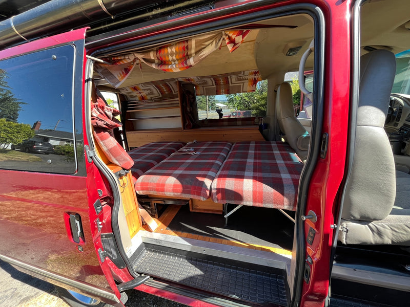 Experience comfort in this RV rental! View of a red camper van's interior with a custom plaid bed and storage. — photo 9
