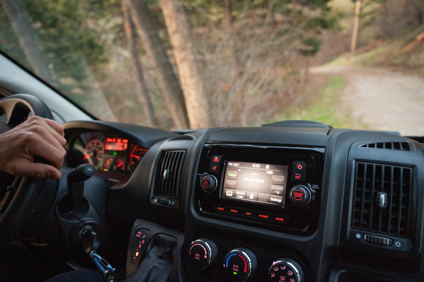 Driving an RV rental: hand on the wheel, dashboard controls and media display visible, with a scenic dirt road and trees outside.