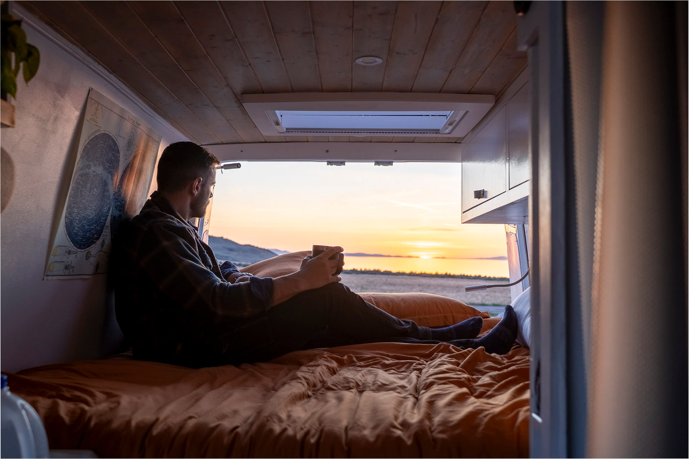 Enjoy stunning sunset views from a cozy RV rental! Man relaxes on the bed of his camper van, looking out at a lake and hills.