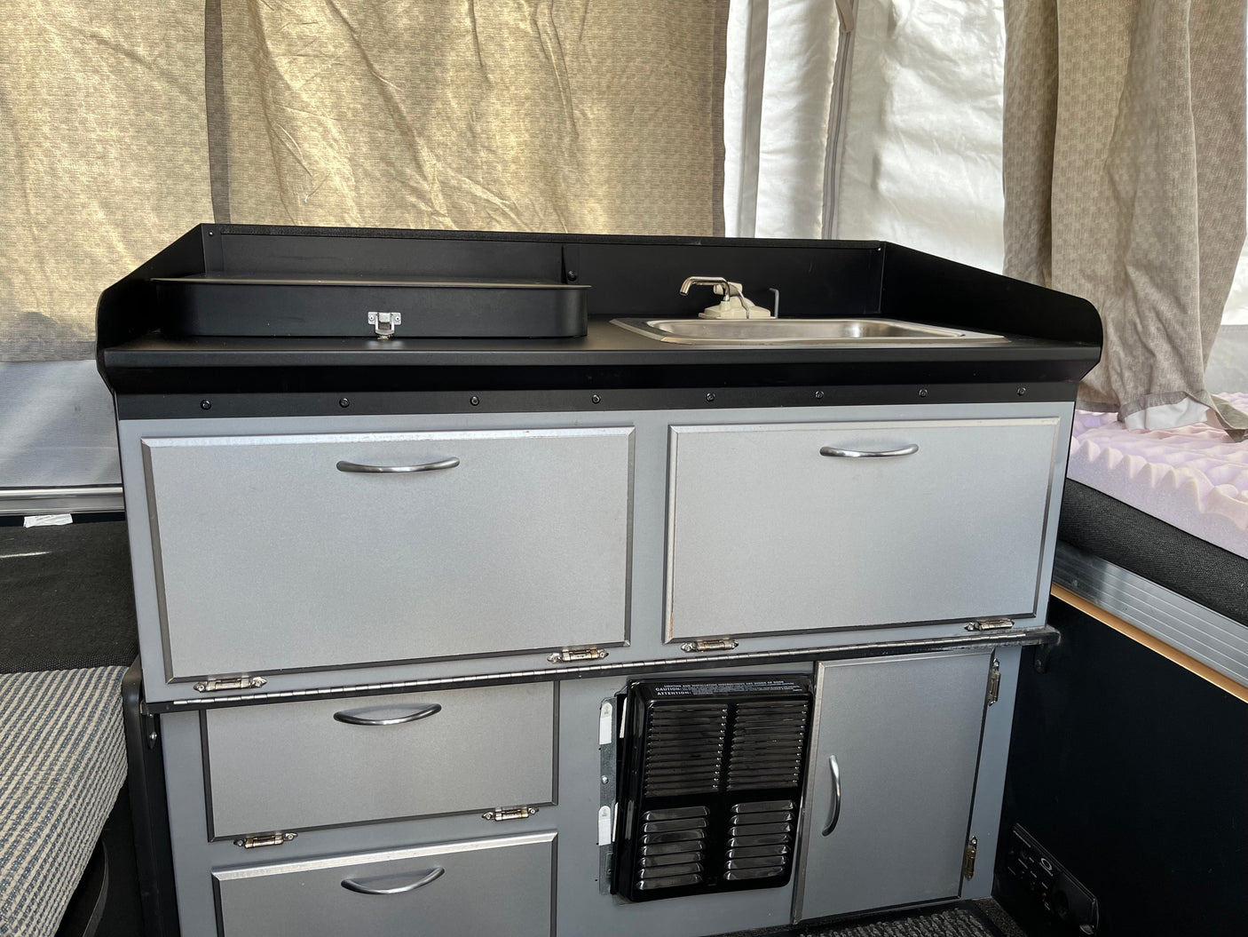 View of a light gray RV rental kitchenette with black counters, sink, faucet, & numerous storage cabinets.