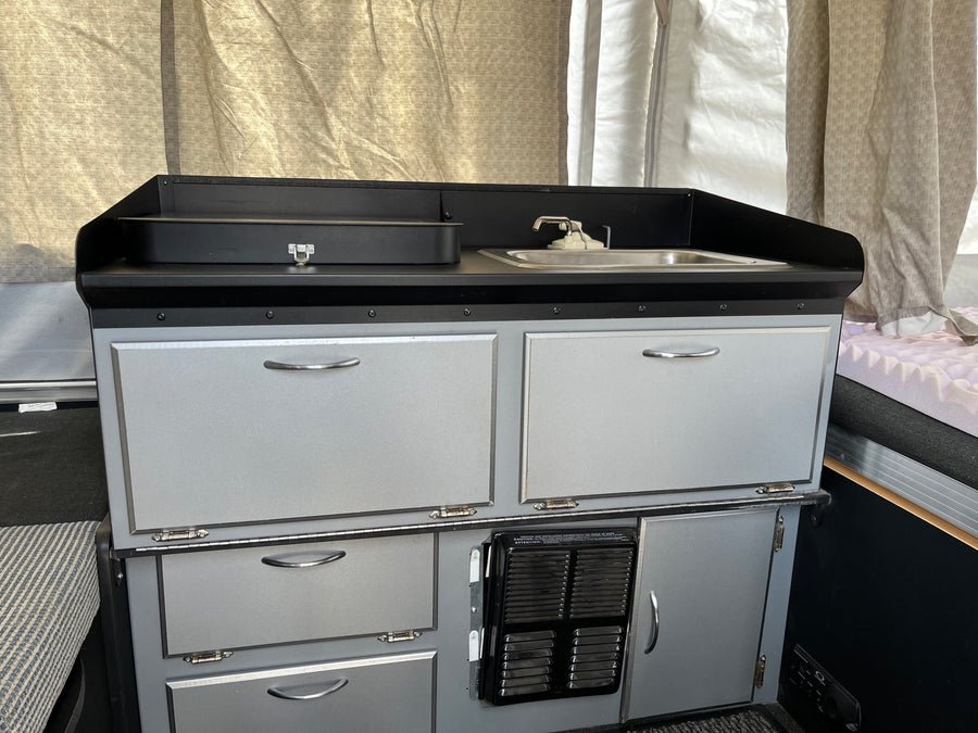 View of a light gray RV rental kitchenette with black counters, sink, faucet, & numerous storage cabinets.