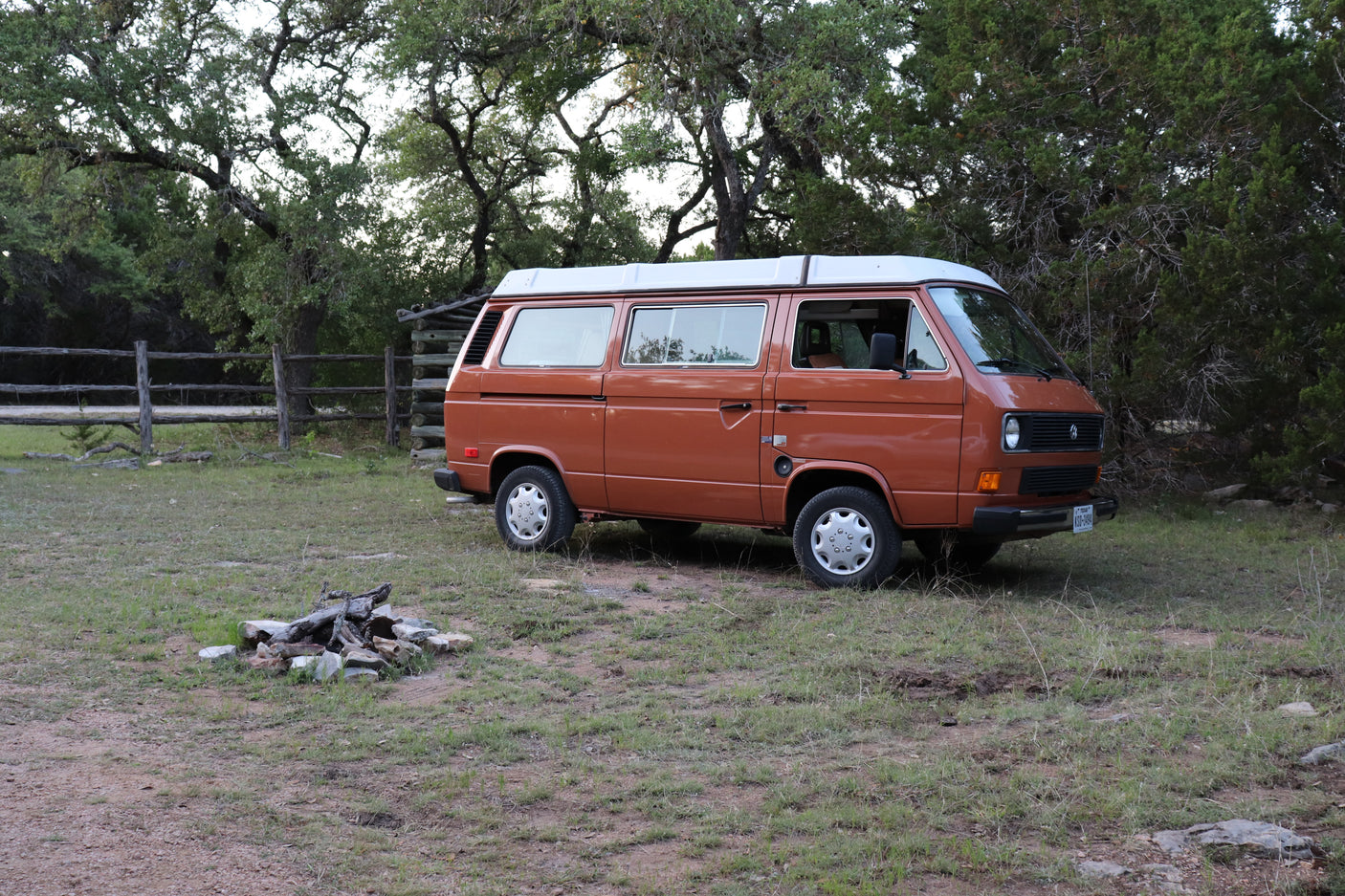 Orange VW camper van, an RV rental, at a rustic campsite with a fire pit, surrounded by lush trees.