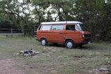 Orange VW camper van, an RV rental, at a rustic campsite with a fire pit, surrounded by lush trees.
