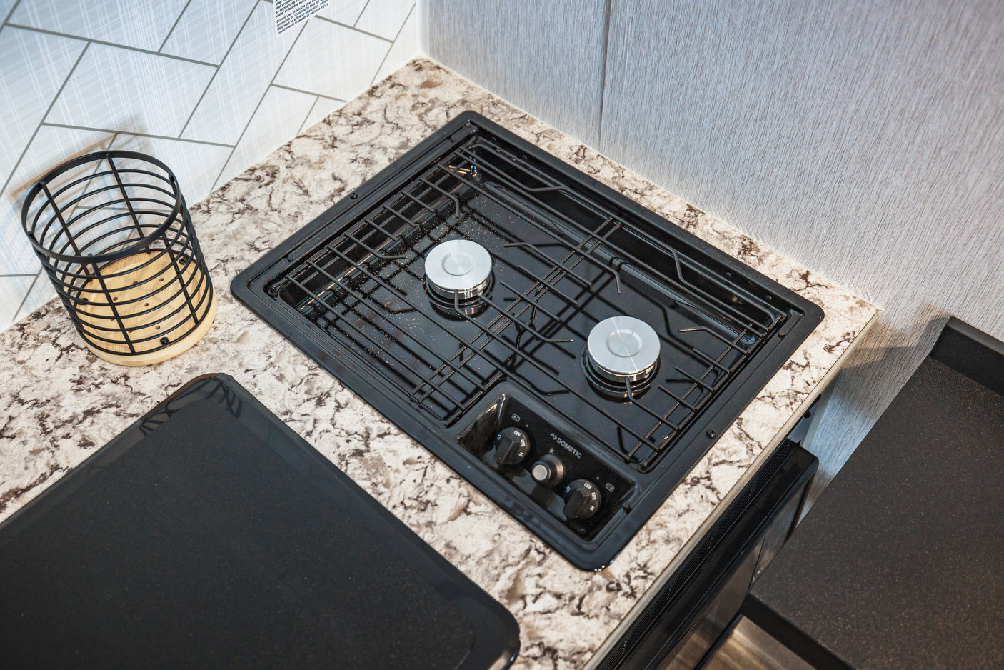 Compact RV rental kitchen with a Dometic two-burner gas stove, speckled countertop, and a light-colored geometric backsplash.