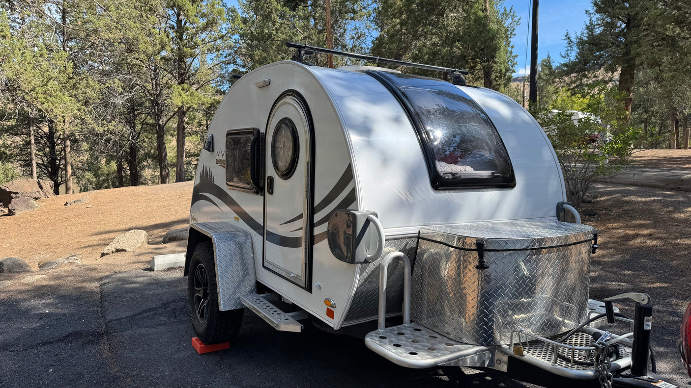 White teardrop RV rental, a Boondock model, parked at a sunny campsite amidst pine trees.