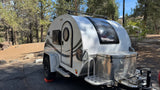 White teardrop RV rental, a Boondock model, parked at a sunny campsite amidst pine trees.