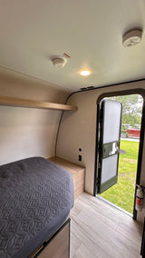 RV rental interior: cozy bed, wooden shelf, smoke alarms, fire extinguisher by the open door to the outdoors.