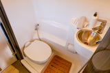 Compact RV rental bathroom: toilet, metal sink, bidet sprayer, and wooden floor mat.