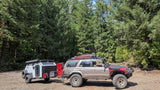 Off-road SUV towing an overland camping trailer through a dense forest. A rugged RV rental option for adventure travel.