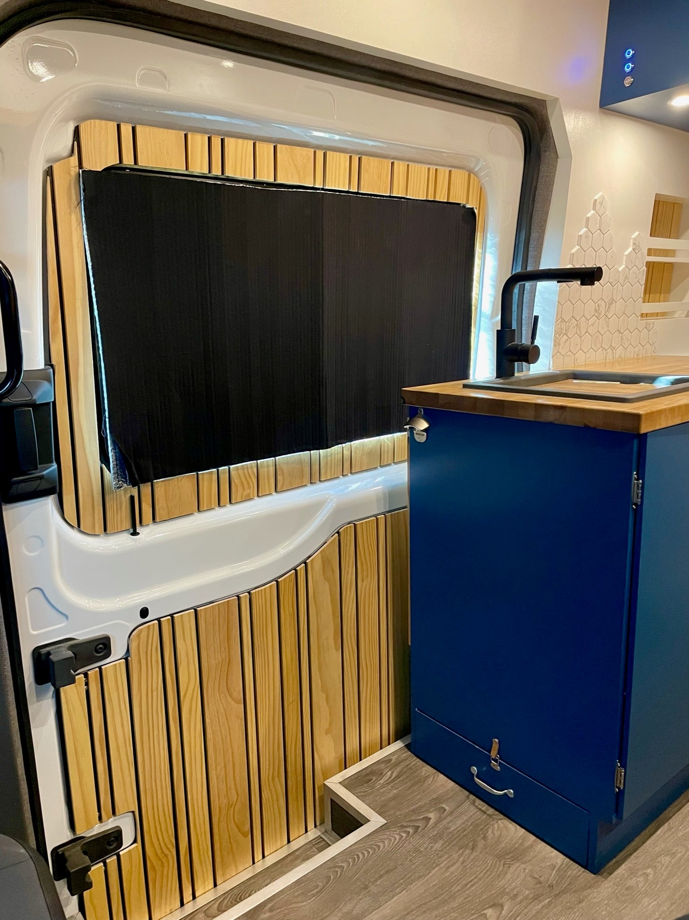 Inside a modern RV rental: custom wood-paneled door, black window blind, and blue kitchen unit with wood counter and sink.