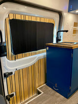 Inside a modern RV rental: custom wood-paneled door, black window blind, and blue kitchen unit with wood counter and sink.