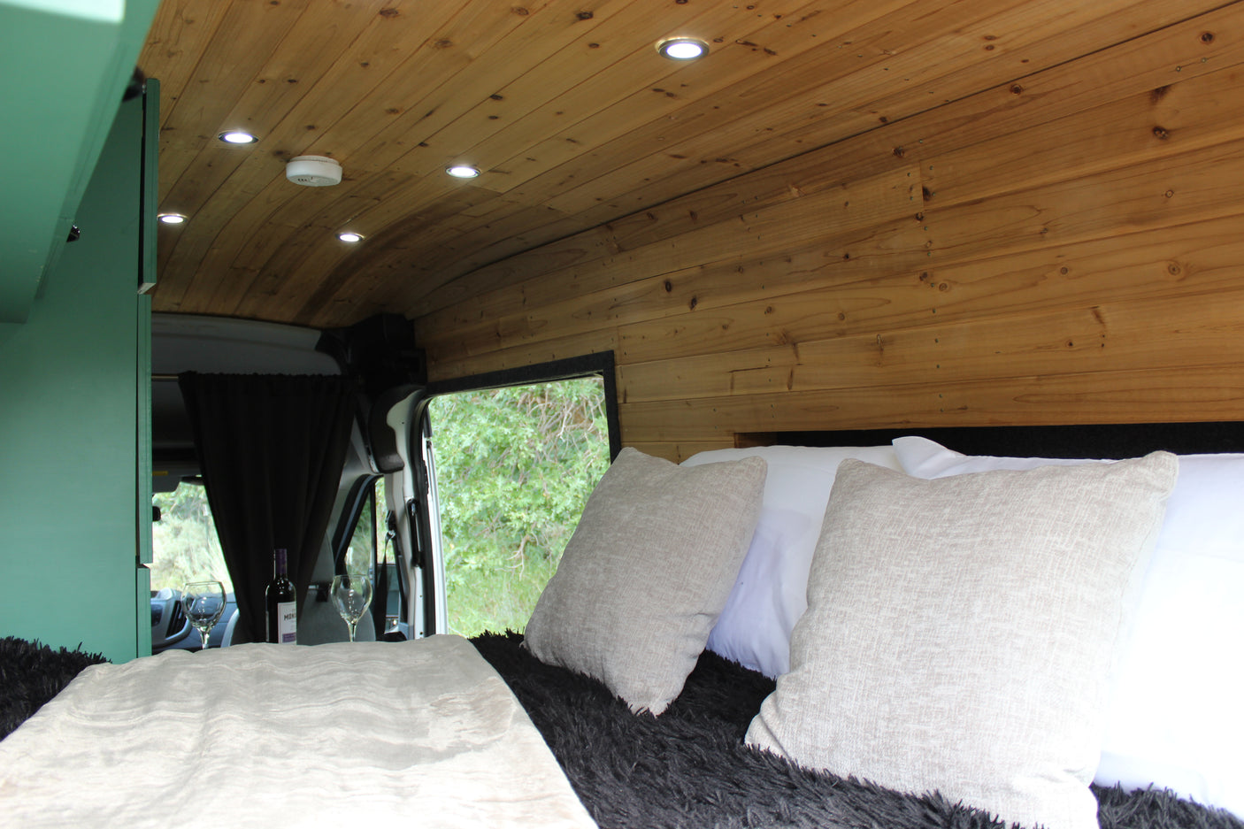 Warm RV rental bedroom: curved wooden ceiling, LED lights, plush bed, and a tranquil nature view. Ready for relaxation.