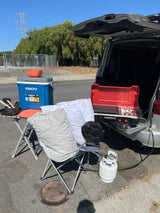 Compact outdoor cooking and relaxation setup beside an RV rental, with a stove, cooler, fan, and chair.