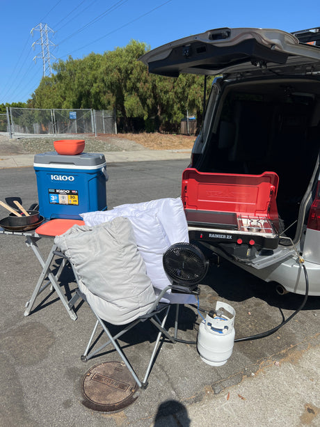 Compact outdoor cooking and relaxation setup beside an RV rental, with a stove, cooler, fan, and chair.