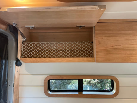 Custom wood interior of an RV rental with an open overhead cabinet, storage net, and small window, ready for travel.