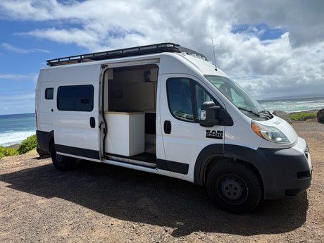 White Ram 2500 rv rental camper van parked on a dirt road by the ocean, its side door open, revealing a minimalist interior.