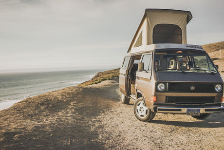 Coastal RV rental dream: Brown Westfalia camper van with pop-top open on a dirt road overlooking the vast ocean.