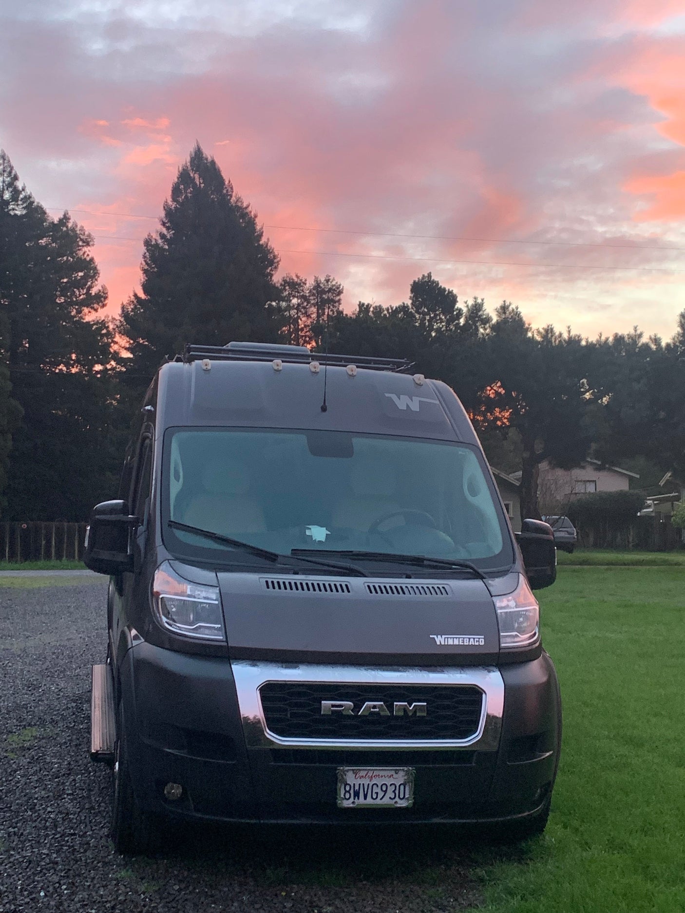 Dark gray Winnebago RV rental van parked at sunset with vibrant pink and orange clouds overhead. Gravel and grass visible.