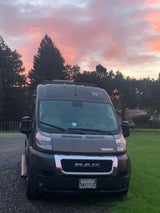 Dark gray Winnebago RV rental van parked at sunset with vibrant pink and orange clouds overhead. Gravel and grass visible.