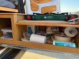 Efficient RV rental kitchen setup: a pull-out drawer with a portable stove, cooking tools, paper goods, and cleaner.
