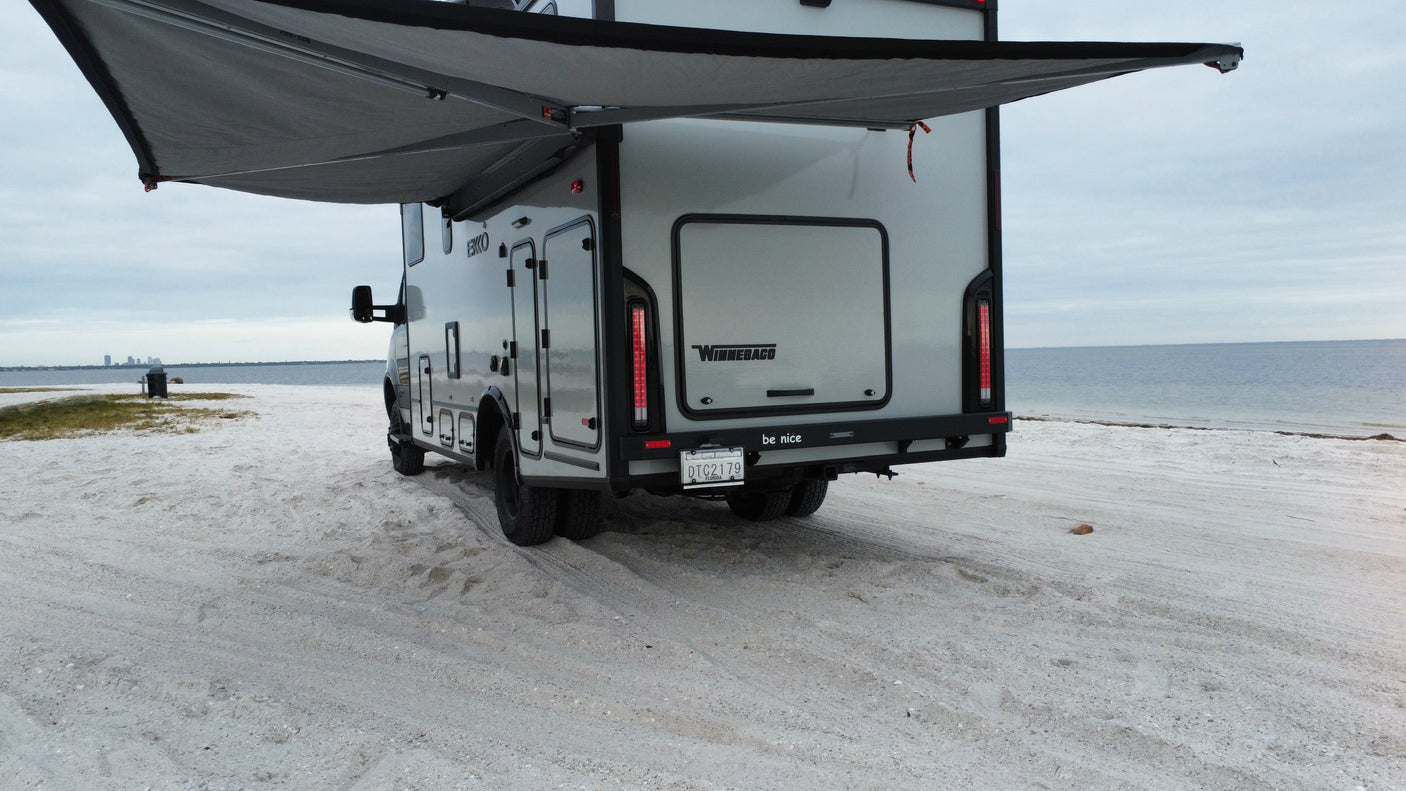 Winnebago Ekko **rv rental** with awning extended on a sandy shoreline, facing the calm ocean & distant city. Great outdoor travel.