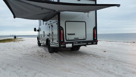 Winnebago Ekko **rv rental** with awning extended on a sandy shoreline, facing the calm ocean & distant city. Great outdoor travel.