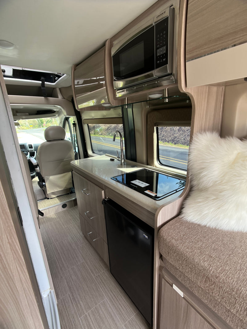 Inside an RV rental, a modern kitchen: microwave, sink, stovetop, fridge, and seating area. Front cab visible beyond. — photo 6
