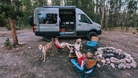 Family enjoying their RV rental: Kids relaxing by a campfire with their dog in a serene forest campsite.