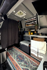 Cozy RV rental interior: compact kitchen area, Dometic fridge, organized overhead food storage, and a vibrant patterned rug.