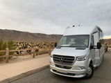 An Airstream rv rental parked roadside near a Cholla Cactus Garden with desert mountains under a cloudy sky.
